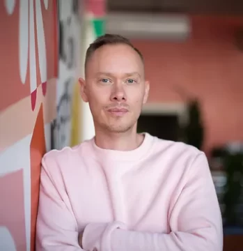 Photograph of Zeke Caligiuri, a white man with short blond hair cut in a fade. In this picture he's wearing a white shirt and leaning against a decorated wall with his arms folded.