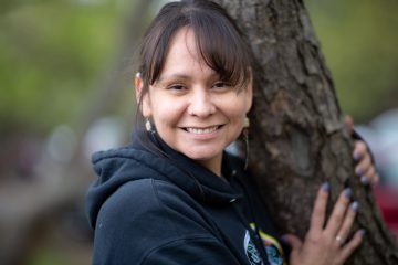 Louise Waakaa'igan is an Ojibwe woman; in this photo, she has mid-length brown hair, a dark blue hoodie, and is touching a tree.