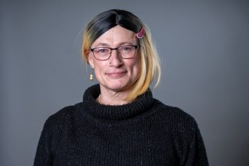 Photo of Davi Gray, a white trans nonbinary person. In this photo, she has shoulder-length blonde hair, pulled behind her right ear, and wears glasses and a black sweater with a rolled neck. (photo credit rebecca smith speak mpls)