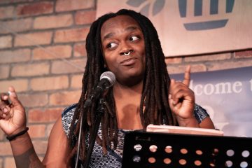 A photo of Taiwana Shambley, a dark-skinned Black woman with chest-length braids. In the picture, she stands at a podium in front of a brick wall, at a microphone, and is holding up her left index finger as she looks at someone in front and to the side of her.