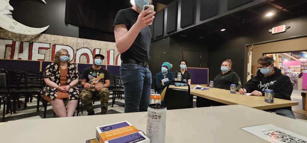 Photo of an attendee at Better Things June 2025 standing and reading their poem from their phone.