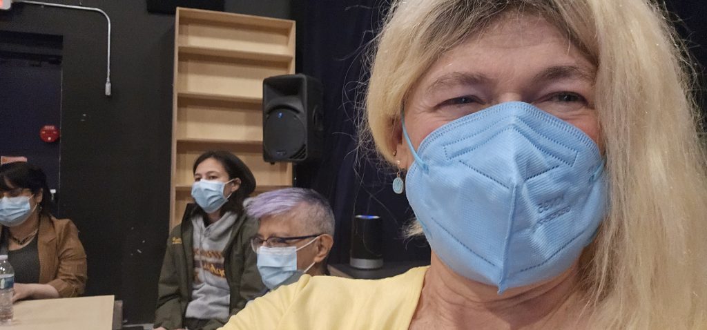Selfie of Davi Gray at Better Things June 2025; in this photo, she is wearing a yellow shirt, a bright blue mask, and shoulder-length blonde hair pulled to one side. Behind her, a couple other attendees sit at the table.
