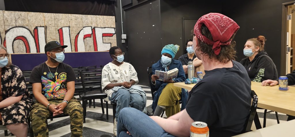 Photo of Better Things June 2025 attendees watching Vanessa - a young Black woman wearing a blue hat - reading a poem from her notebook.