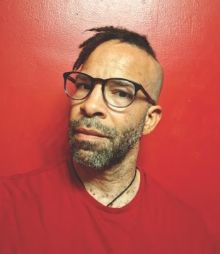 Photograph of Douglas Kearney, a Black man with light brown skin; he's wearing a red shirt, a necklace, and glasses, and has short facial hair and a partially shaved head with short braids flipped to one side; he stands in front of a red background. His expression is serious and inquisitive.