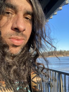 Photograph of Lamar Renville, a Dakota two-spirit poet with long brown hair and brown skin, standing near a lake. On the far shore, mixed evergreen and leafless deciduous trees.