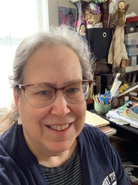 Photograph of Nell Ubbelohde, a white woman elder with long white hair pulled back. She's wearing glasses, a dark blue shirt, and a wide smile. In the background is a desk with lots of papers and pens.