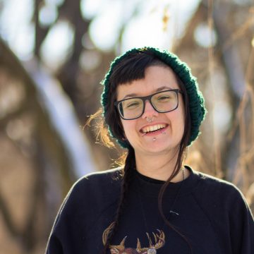Photograph of Ollie Schminkey, a smiling white nonbinary poet with shoulder-length hair tucked under a green knit hat; they are wearing large glasses and a brown sweatshirt and are posed in front of a blurred background of trees. (Photo credit Mel Nigro)