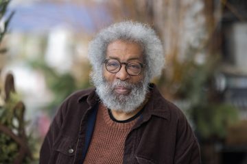 Photograph of David Lawrence Grant, a Black man with silvered afro, beard, and mustache, wearing a dark brown jacket over a lighter brown shirt.