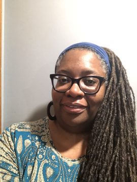 Photograph of Sherrie Fernandez-Williams, a Black woman with glasses and very long dreadlocs. In this photo she is wearing a blue headband and patterned blue-and-gold shirt.