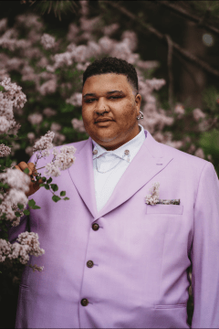 Photograph of Kanoro Day, a Black man with short curls on top of his head and short sides; in this photo, he's wearing a pink suit jacket over a white shirt and standing next to a plethora of flowers.