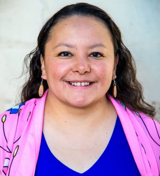 Photograph of Oogie Push, a Native woman with long, wavy brown hair; in this photo, she's wearing earrings and a pink jacket over a blue shirt.