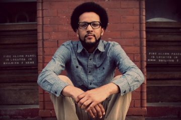 Photograph of Chaun Webster, a Black man with light skin; in this photo, he has a mid-length afro, glasses and trimmed facial hair, is wearing a denim shirt, and is seated in front of a brick wall.