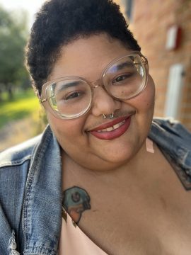 Photograph of TaneshaNicole, a Black nonbinary person with light brown skin, short curly hair, and a light mustache. In this photo they are wearing big glasses, a septum ring, and a denim jacket.