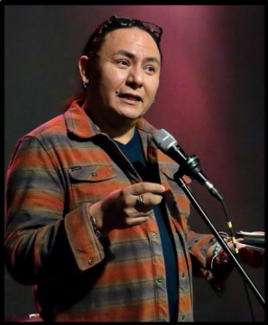 Photograph of Nick Metcalf, a two-spirit American Indian. In this photo, they are wearing a striped collared shirt over a t-shirt and standing in front of a microphone with one hand raised.