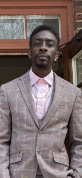 Photograph of Coley Gates, a Black man with dark skin; in this photo, he is wearing a pink collared shirt under a suit jacket.
