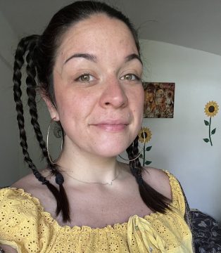 Photograph of Nana J, a white woman with dark brown hair; in this photo she wears braids and a yellow dress and is almost smiling.