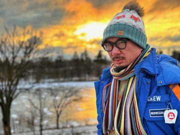 Photograph of Bao Phi, an Asian-American man with a black goatee and mustache. In this photo, he is standing in a wintry landscape with an orange sunlit cloudscape; he wears a blue coat, multicolored scarf, sweater, and hat, and black-framed plastic eyeglasses. His expression is fierce.
