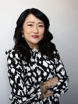 Photograph of Sun Yung Shin, a Korean-American woman with long black hair and freckled cheeks. In this photo she is wearing a black and white mottled blouse, bracelets, large ring earrings, and a subdued friendly expression.