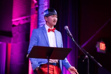 Photograph of Fong L., a Hmong-American man with a light mustache. In this photo, he is wearing red pants, a white shirt, red bow tie, and blue plaid jacket, with a paneled blue hat. He stands before a microphone.