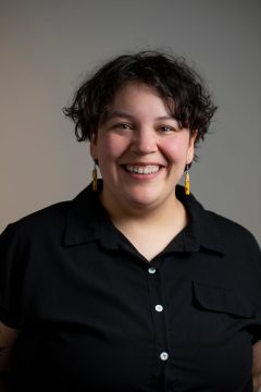 Photograph of Tyra Payer, a Native nonbinary person with short dark hair. In this photo they are wearing yellow earrings, a black button-down shirt, and a wide smile.