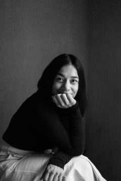 Photograph of Ari Tison (Bribri), an Indigenous woman with light brown skin. In this photo she is wearing long black straight hair, a long-sleeved black shirt, and white pants; she is seated and leaning forward, right elbow on knee, resting her chin on her right palm, half-concealing a half-smile.