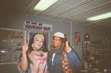 Photograph of BakiBakiBaki, an Afro-indigenous person with medium-brown skin. In this photo they are wearing a jean jacket and a particolor vest, long twining braided hair, a knit white cap, and a wide smile; they are holding up index and middle fingers of their left hand.