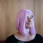 Photograph of Davi Gray, a white trans nonbinary woman with gray eyes. In this picture she is wearing pink hair, a jeweled stud in her right nostril, and a black shirt. She's standing in front of a blurred wood background and staring off to camera right.