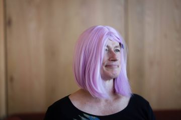 Photograph of Davi Gray, a white trans nonbinary woman with gray eyes. In this picture she is wearing pink hair, a jeweled stud in her right nostril, and a black shirt. She's standing in front of a blurred wood background and staring off to camera right.