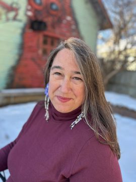 Photograph of Heid E. Erdrich, a light-skinned Ojibwe woman. In this photo, taken outdoors in front of a blurry winter background, she stares at the camera, and wears long reddish, brown, and white hair; a maroon turtleneck; long beaded earrings; and a slight smile.