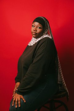 Photograph of Isha Camara, a dark-skinned Black woman. In this photo she is wearing a black top, dark denim pants, and a patterned black-and-white hijab. She is seated in front of a red background. Her eyes are surrounded by colored stars and she has a serious expression, looking directly at the camera.