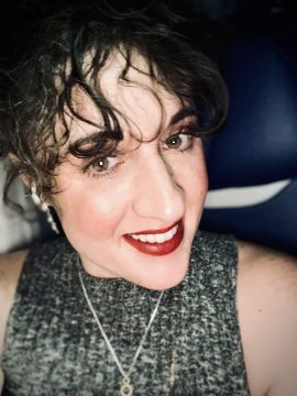 Photograph of Cecily McMillan, a Latine person with light skin and dark brown hair. In this picture she's wearing a sweater vest, earrings and red lipstick, and shows her teeth in not quite a smile.