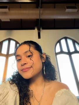 Photograph of Isavela Lopez, a Latina woman with light brown skin and very dark hair. In this photo, she's wearing a white top with puffy sleeves, two necklaces, hand-crafted pendant earrings, a nose ring, and long wavy hair pulled to the back and side.