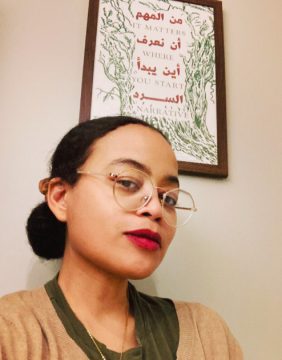Photograph of Sagirah Shahid, a Black woman with light brown skin. In this photo, her hair is pulled back into a low bun; she's wearing thin gold framed glasses, a tan sweater over olive shirt, a gold necklace, and a skeptical expression. Behind her on the wall, a framed print with Arabic writing and an English phrase, "IT MATTERS WHERE YOU START - A NARRATIVE."