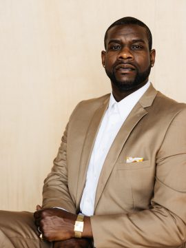 Photograph of Lester Batiste, a Black man with dark skin, very short hair, and a trim beard and mustache. In this picture he's wearing a tan suit with pocket square, white shirt, gold watch, stud earrings, and two rings, and he's got a serious expression as he looks into the camera.
