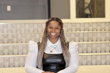 Photograph of Lt. Sunnie, a Black woman with medium brown skin and very long braided hair descending from both sides. In this photo she's wearing a leather-look top over white collared shirt, silver necklace, large earrings, and a huge smile.