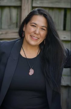 Photograph of Rosetta Peters, a Dakota woman with medium brown skin and long black hair. In this picture, she is wearing a navy blazer over a navy shirt, a sliced-stone pendant, and pendant earrings; in the background is a weathered wooden fence.