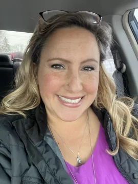 Photograph of Suzanne Locke, a white woman with long blond hair. In this photo she's wearing a black coat over a fuchsia top, two necklaces, a nose stud, and glasses propped on top of her head. She's got a wide smile and freckles.