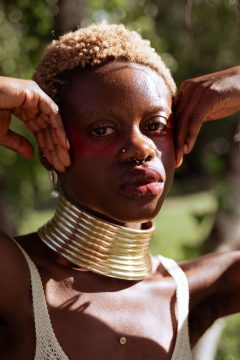 Photograph of Ashe Jaafaru, a Black woman with dark skin, wide eyes, and full lips; in this photo she's wearing short, curly, two-toned hair, nose piercings, earrings, and a gold multi-ring collar. She stares at the camera with a calm expression.