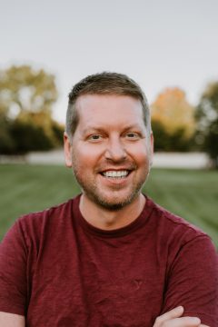 Photograph of Ben Westlie, a young white man with dark blond hair. In this photo, he wears short hair, a short scruff of beard and mustache, a wide smile, and a crimson velour T-shirt. He's standing, arms crossed, in front of an out-of-focus grassy field.