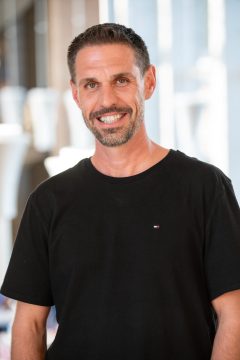 Photograph of Andre Anderson, a white man with dark brown hair. In this photo he is wearing a wide smile, short hair, a short beard and mustache with sprinkles of gray, and a black T-shirt. He stands, arms slightly akimbo, in front of a blurred background.