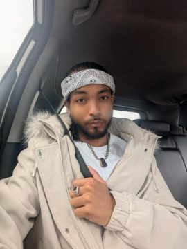 Photograph of Armand McCoy, a young Black man with medium-brown skin, short hair, and a short beard and mustache. In this photo he's wearing a light tan coat over a white T-shirt, a bandanna around his forehead, a ring on his middle finger, and a necklace. He's sitting in a vehicle and has a neutral expression.