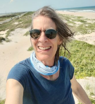 Photograph of Jennifer Landretti, a white woman with medium-short graying brown hair. In this photo, she's wearing a blue sleeveless top, a blue gaiter around her neck, and dark sunglasses, and is standing in dunes behind an ocean shore.