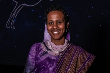 Photograph of Sabrin Nur, a young Black person with dark skin and short black hair. In this photo, they have a wide smile and are wearing pearl necklaces and earrings and a purple garment with a purple head covering and a striped cloth over their left shoulder. Behind them are what look like blue stars on a black background, with a constellation tracing barely visible.