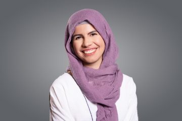 Photograph of Sana Wazwaz, a young Palestinian American woman with light brown skin. In this photo, she is wearing a purple hijab and a white top, smiling at the camera against a gray background.