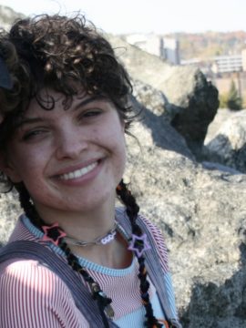 Photograph of Adrien Wright, a nonbinary white person with dark brown hair. In this photo, they are wearing blue striped overalls and a red/white and blue/white striped shirt, a safety-pin necklace, and mid-length curly hair with two long pigtails. They're somewhere outside, with a rock formation in the background.