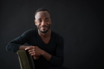 Photograph of Antonio Duke, a young Black man with medium-dark brown skin. In this photo, he's wearing short freshly trimmed hair, a trim goatee and mustache, and a long-sleeve black shirt. He's seated and smiling.