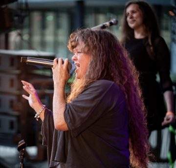 Photograph of Desdamona, a middle-aged white woman with very long wavy brown hair. In this photo, she's on stage singing into a microphone and wearing a black top and a bracelet; behind her, a blurred background including another singer.