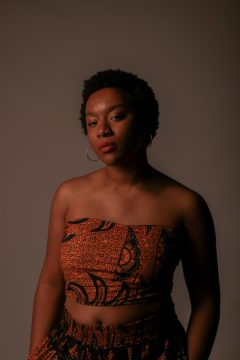Photograph of Eemanna, a young Black woman with medium-dark brown skin. In this photo she's wearing a matched pair of stretch pants and bandeau with orange and black patterning, large hoop earrings, a nostril ring, and short natural hair.