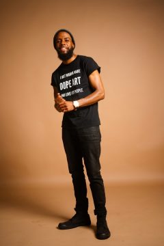 Photograph of Joe Davis, a Black man with medium brown skin and black hair. In this photo, he's wearing a hat, a beard and mustache, and all-black clothes, including a T-shirt which says, "I just wanna make dope art and love my people."