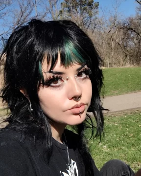 Photograph of Nen G Ramirez, a Chicanx person with light skin and black hair with green splashes. In this photo, they are sitting in a park; their face has multiple piercings and their eyelashes are enormous.